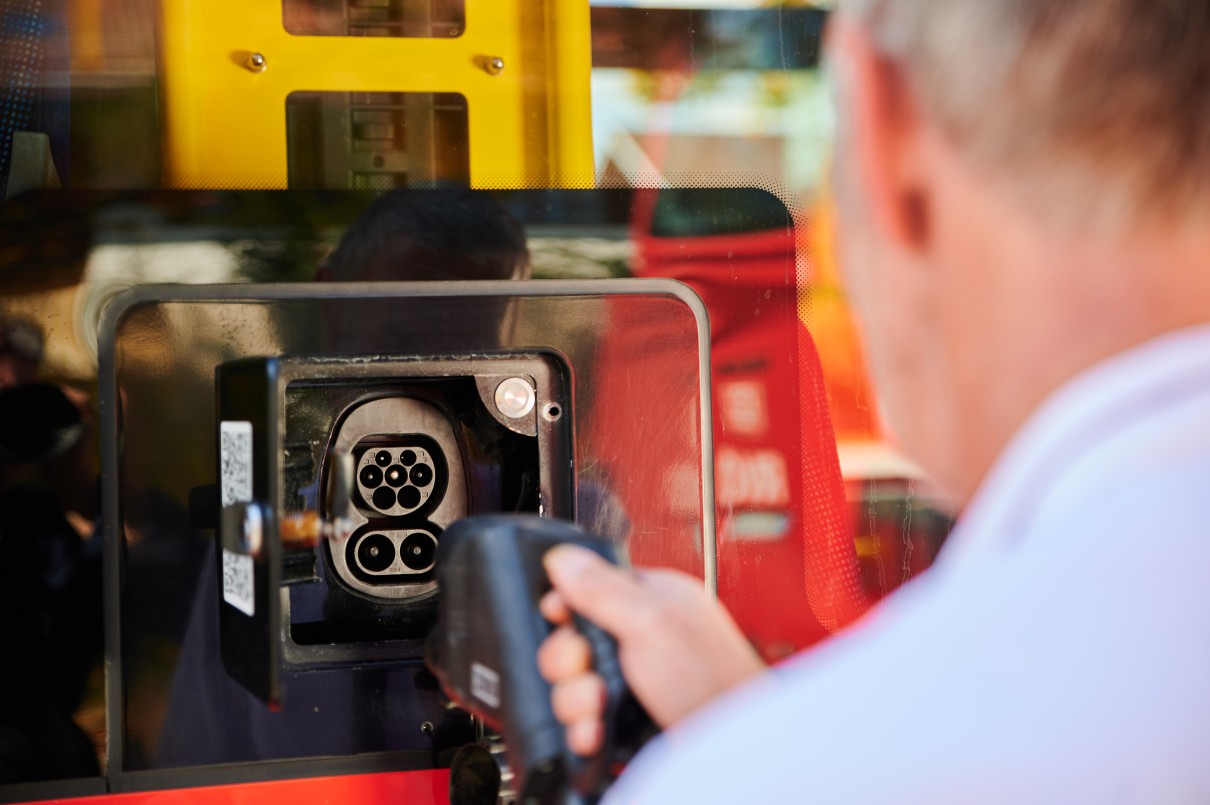 Eine Person hält einen Ladestecker und richtet ihn auf den Ladeanschluss eines Batteriebusses. Der Ladeanschluss und der Stecker sind gut sichtbar im Bild, während die Person nur teilweise von hinten 