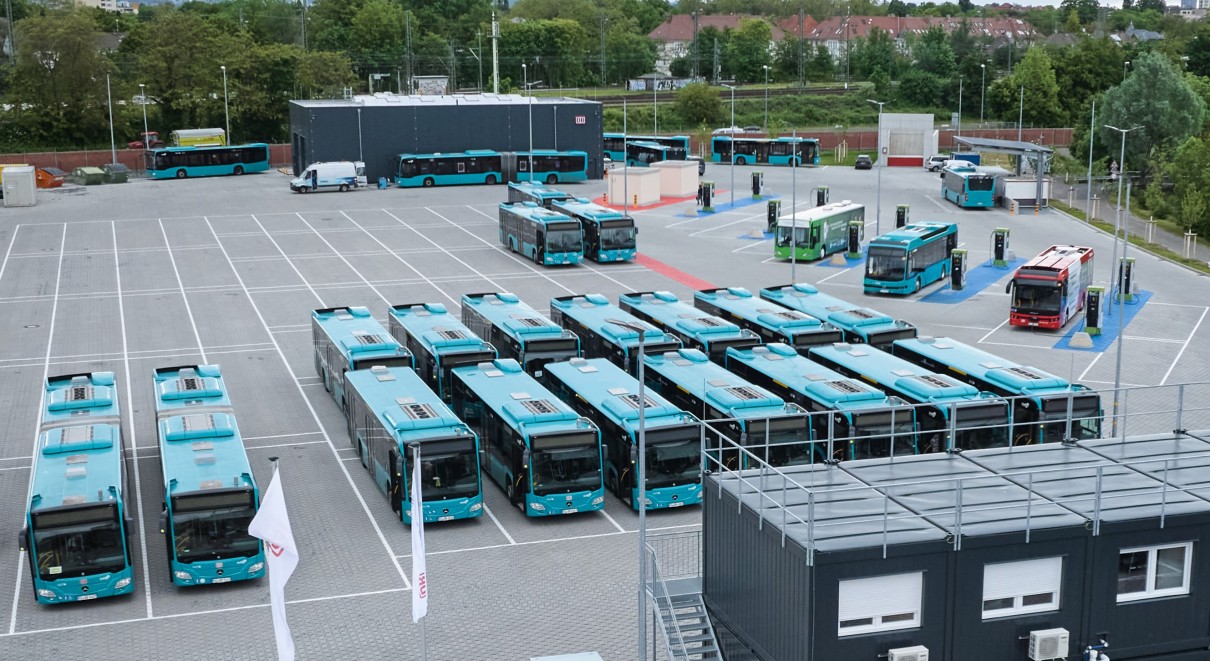 A bus depot with lots of blue buses parked neatly in rows. In the background a green and red bus are connected to charging points. A grey office building and flags are also visible.