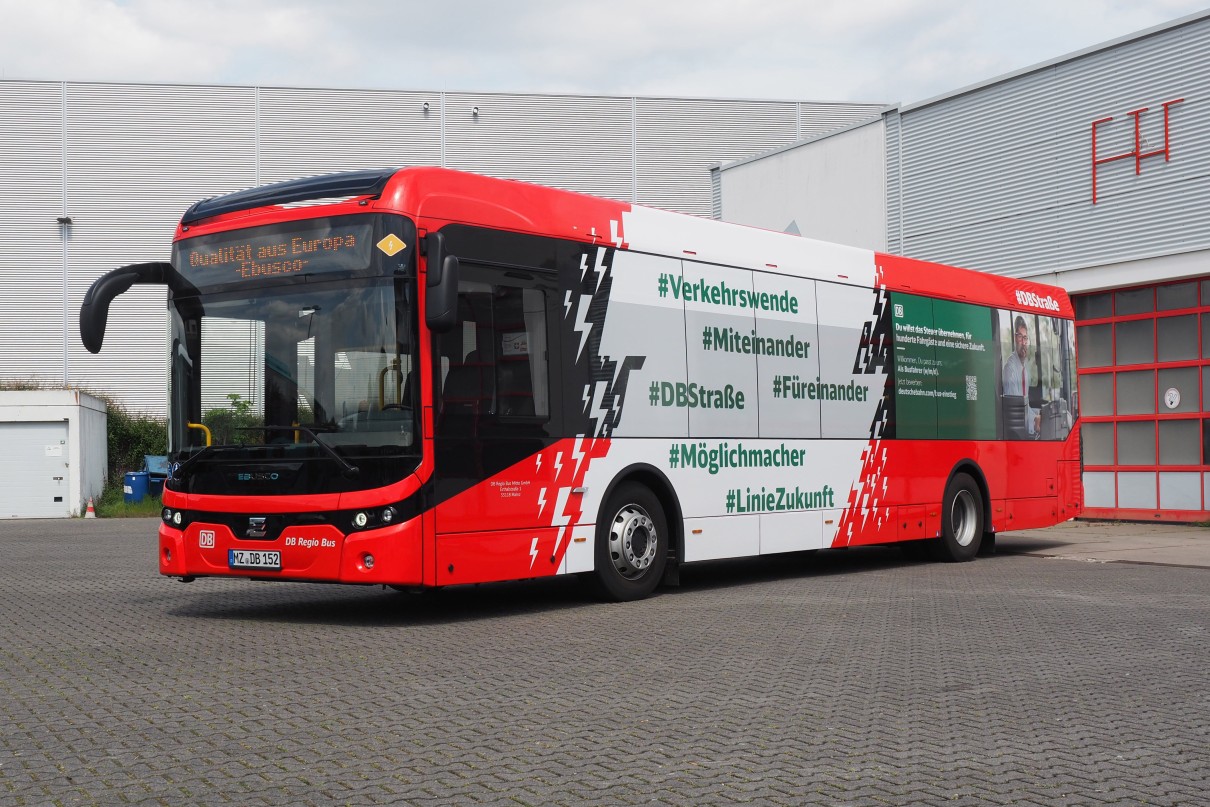 ): The picture shows a red DB Regio Bus electric bus with the inscription “Ebusco”. Several green hashtags such as #Verkehrswende and #LinieZukunft can be seen on the side of the bus. The bus is parke