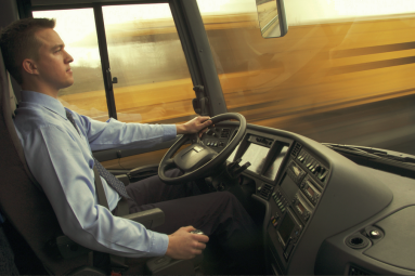 A bus driver in a shirt and tie sits concentrated at the wheel of a bus, as the background rushes by in a blur.